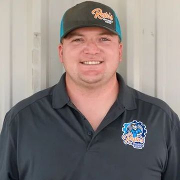 Smiling person in a dark polo shirt and green cap, standing indoors against a light wall.