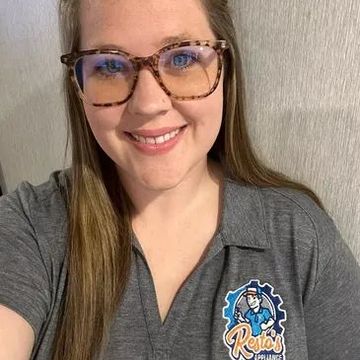 Smiling person wearing glasses and a gray shirt with a sports logo, taking a selfie indoors