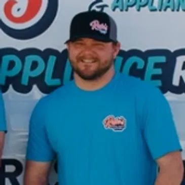 Man in blue T-shirt and black cap smiling in front of a promotional banner