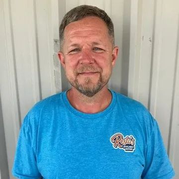 Man in a blue T-shirt standing indoors, smiling at the camera.