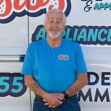 Man in blue shirt standing in front of an appliance service truck, smiling with hands clasped.