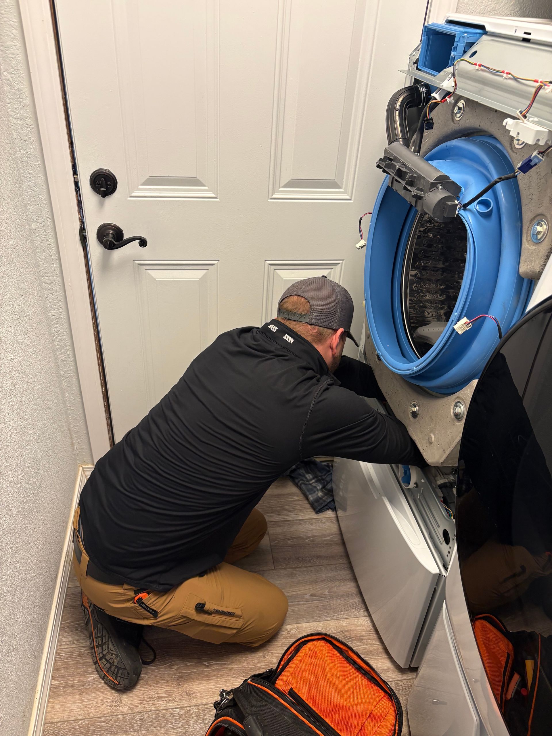 Technician kneels beside a large blue industrial machine, working on its lower panel in a utility room.