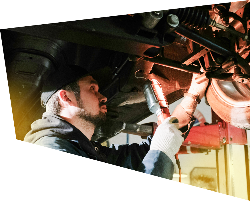 A mechanic in a cap and work gloves uses a portable light to inspect the undercarriage of a car on a lift. | J's Automotive