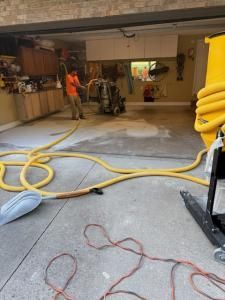 Person using a water extraction machine in a flooded garage; yellow hose and equipment.