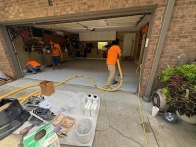 Workers applying a coating to a garage floor; equipment and supplies are in the foreground.