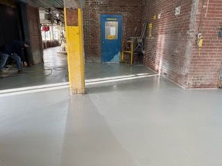 Light gray epoxy floor in industrial setting. Blue door, brick walls, and yellow support column visible.