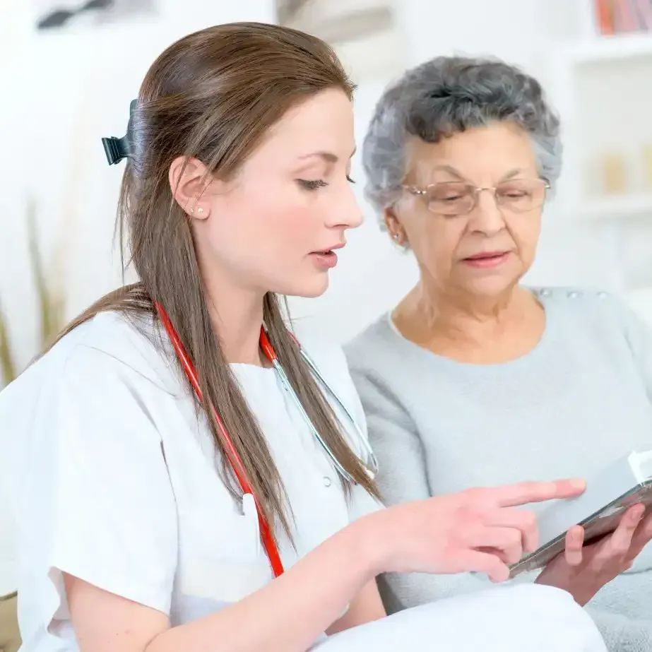 Une infirmière montre une tablette du doigt à un patient âgé, à son domicile.