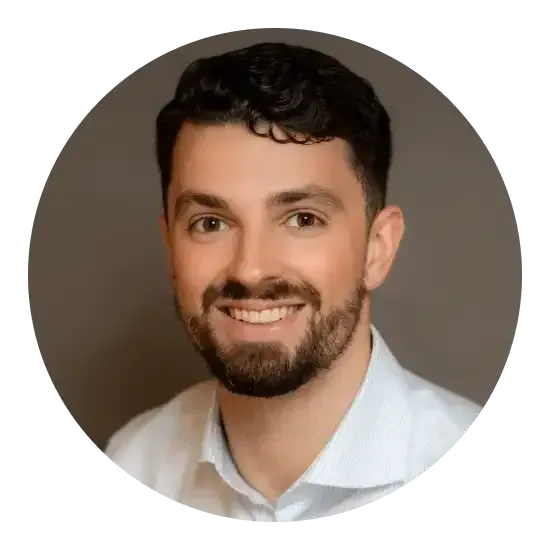 Un homme aux cheveux et à la barbe foncés sourit, vêtu d'une chemise boutonnée bleu clair, devant un fond gris.