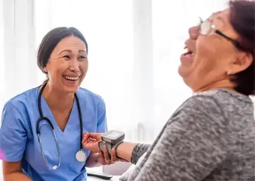 L'infirmière prend la tension du patient. Ils rient tous les deux. La scène se déroule dans un cabinet médical.