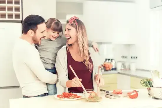La famille cuisine ensemble, riant dans la cuisine : le père tient l'enfant, la mère remue le repas.