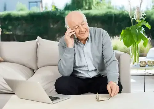 Une personne âgée assise sur un canapé, parlant au téléphone, utilisant un ordinateur portable et souriante.