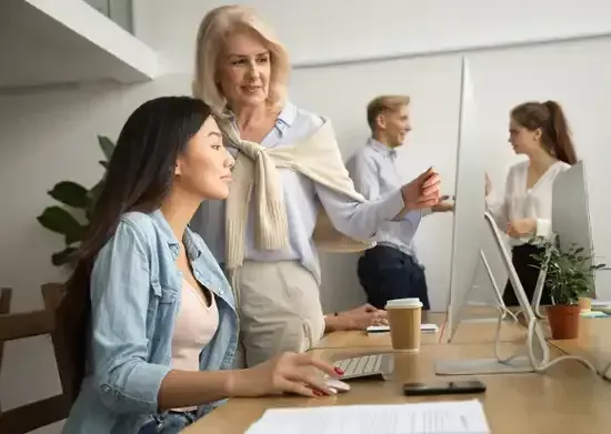 Une femme travaille sur un ordinateur, guidée par un collègue plus âgé présent au bureau ; d'autres collègues se trouvent à proximité.