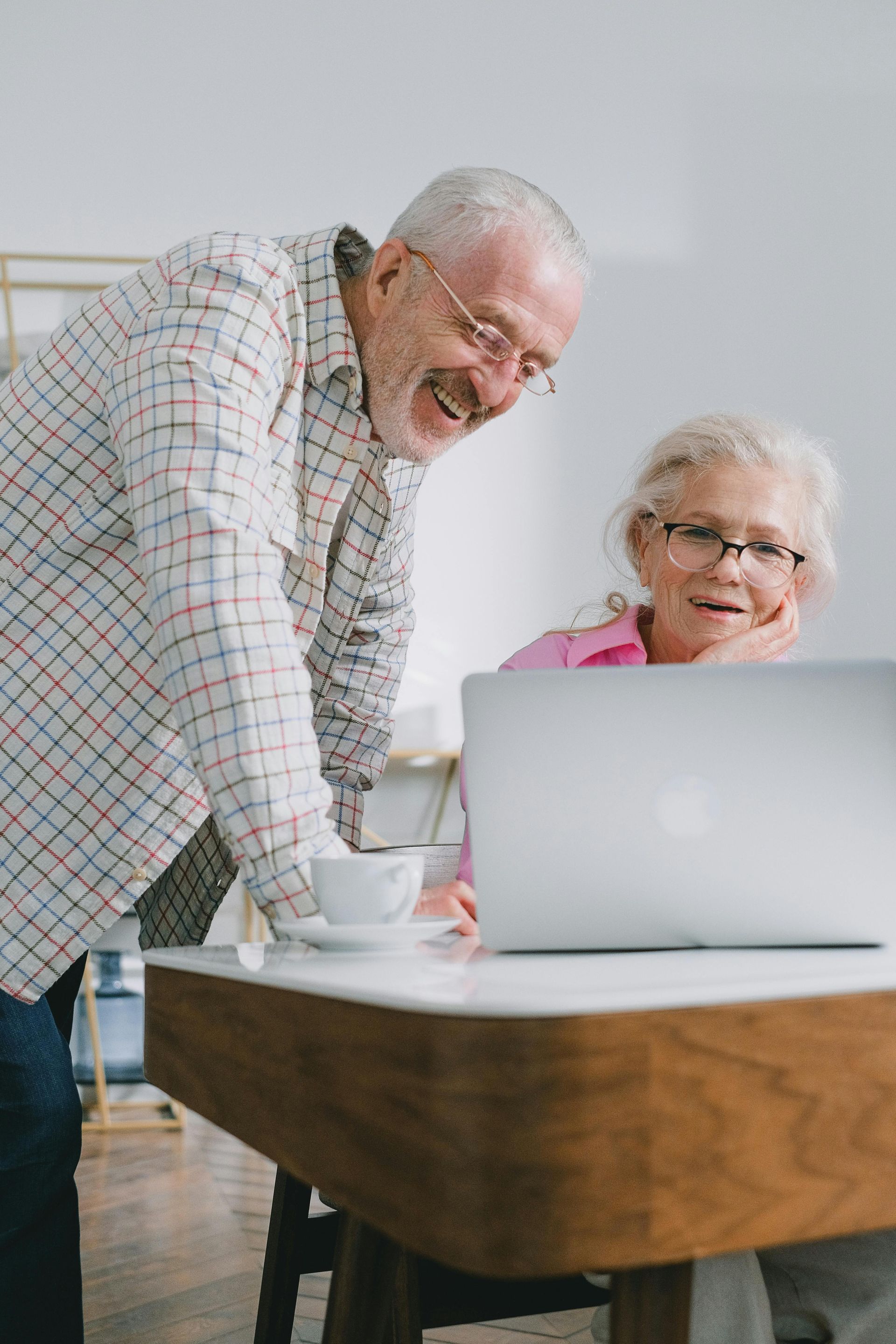 Two people smiling at a laptop beside a wooden table in a bright room
