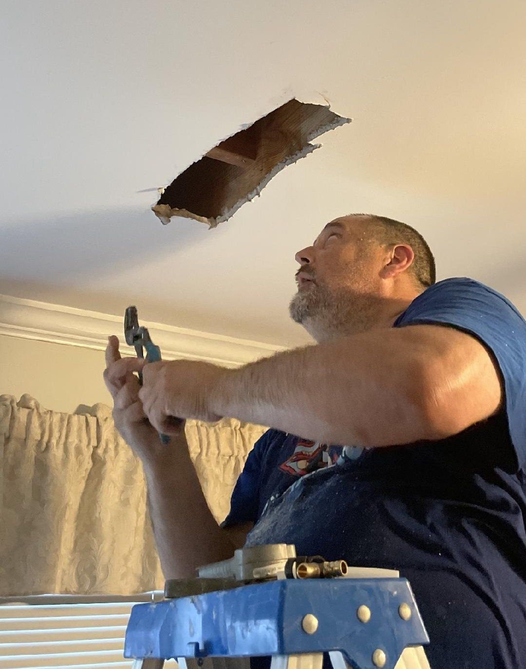 Man on ladder repairing ceiling, holding tools, with a hole above him.