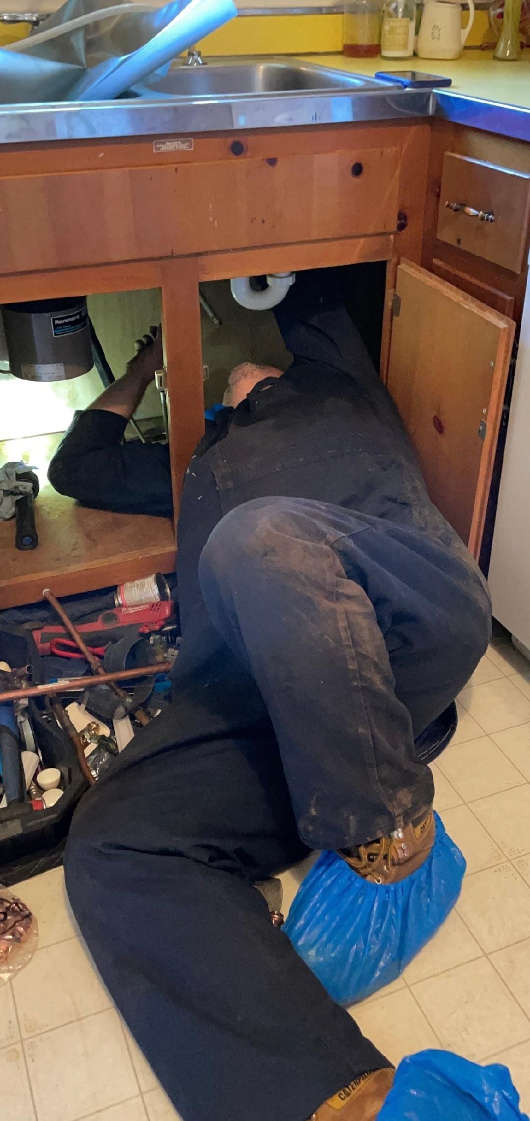 Person working under a kitchen sink. They are wearing blue shoe covers, a black jumpsuit.