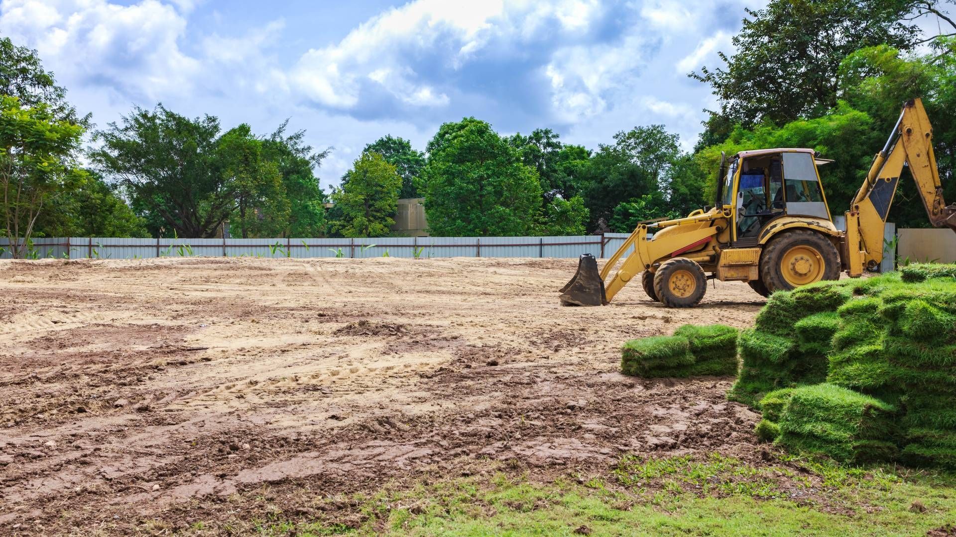 Backhoe on a muddy construction site, stacks of grass, trees, and cloudy sky.