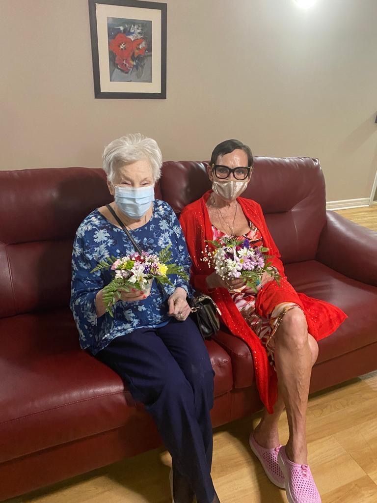 two women wearing face masks sit on a couch holding flowers