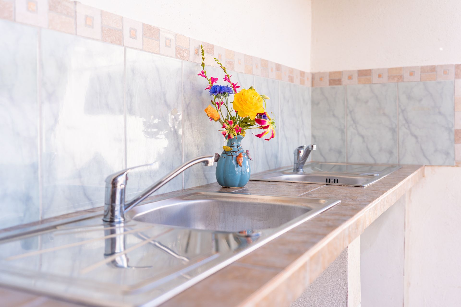 Kitchen with stainless steel sink, stove top, and a vase of colorful flowers against tiled wall.