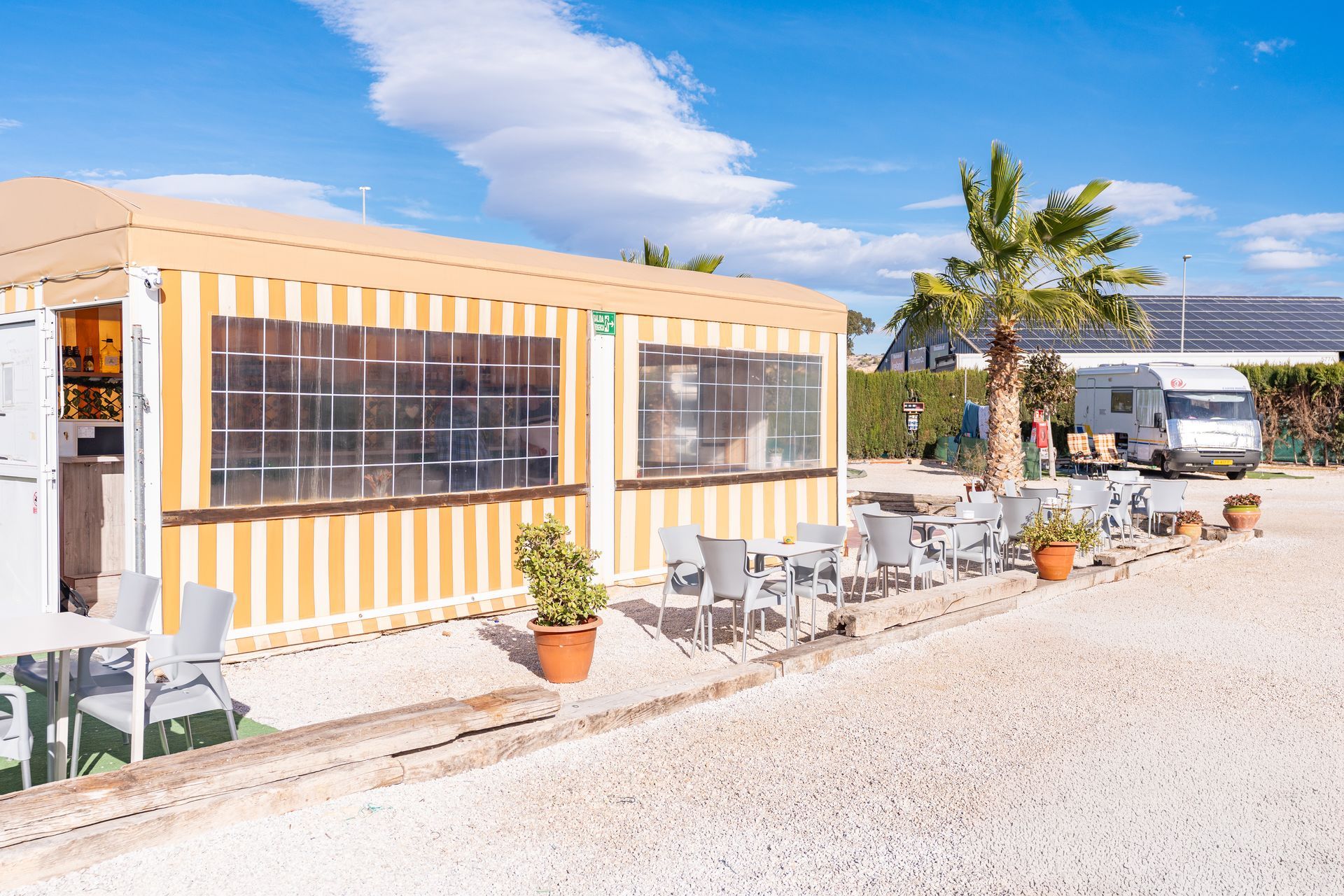 Cafe with outdoor seating, near a palm tree and RV, against a blue sky.