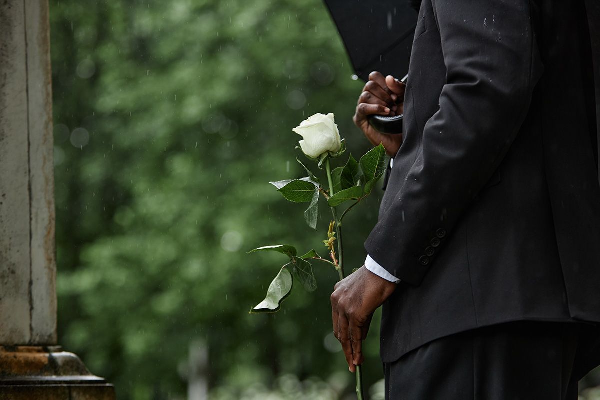 Person in black suit holding a white rose, standing outdoors in the rain.