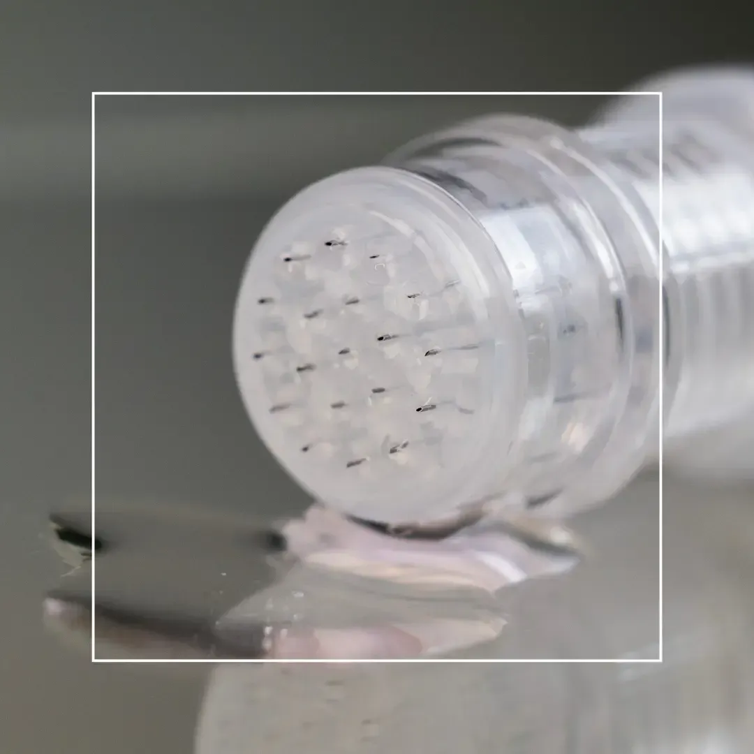 A close-up of a microneedling device tip with tiny needles, positioned above a small drop of clear liquid on a surface.