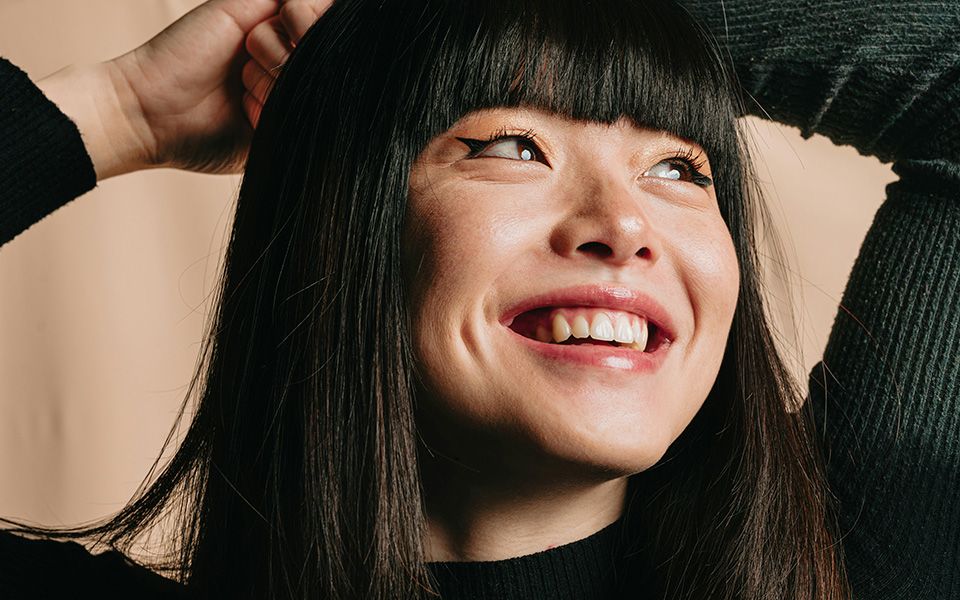 Vrouw glimlacht en kijkt omhoog. Zwart haar met pony, lichte huid, draagt ​​een zwart shirt, handen achter het hoofd.