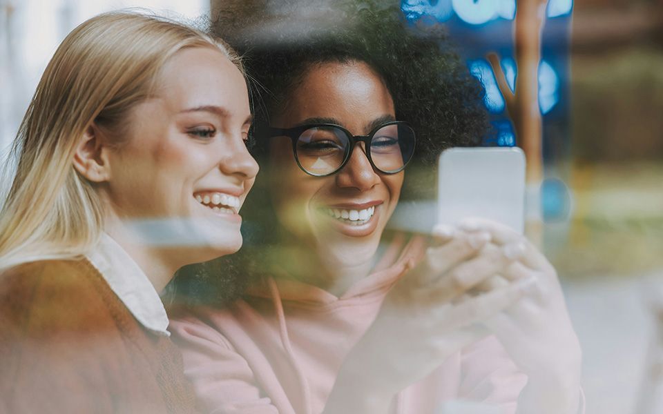 Twee vrouwen glimlachen naar een smartphone, mogelijk makend een selfie, gezien door een raam.