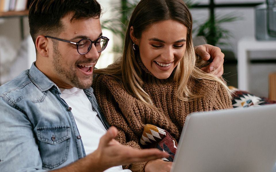 Een lachend stel kijkt binnen naar een laptopscherm. De man gebaart enthousiast.