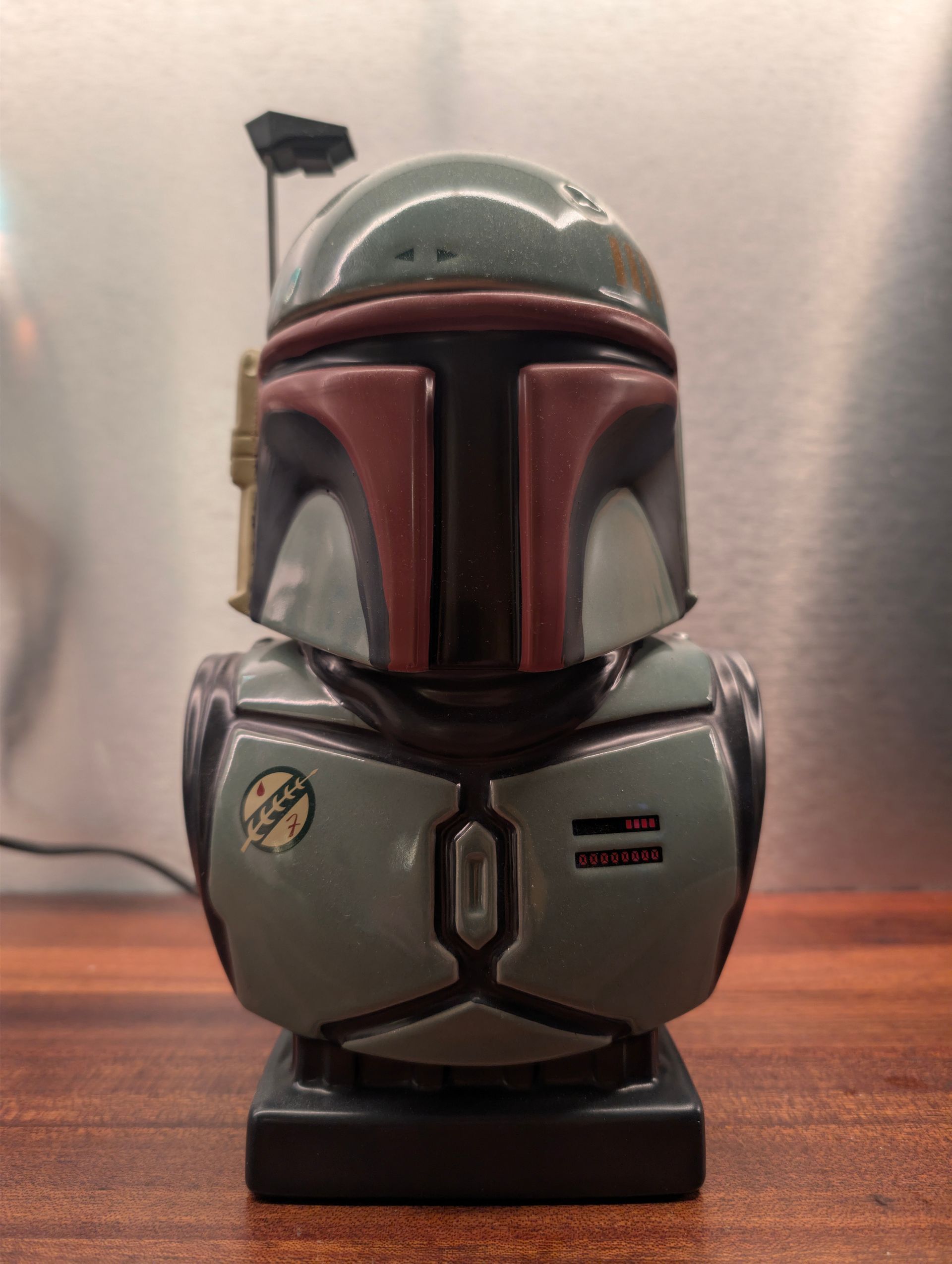 Lamp in de vorm van een Boba Fett-helm, groen, rood en zwart, op een houten voetstuk, met een zwart netsnoer.