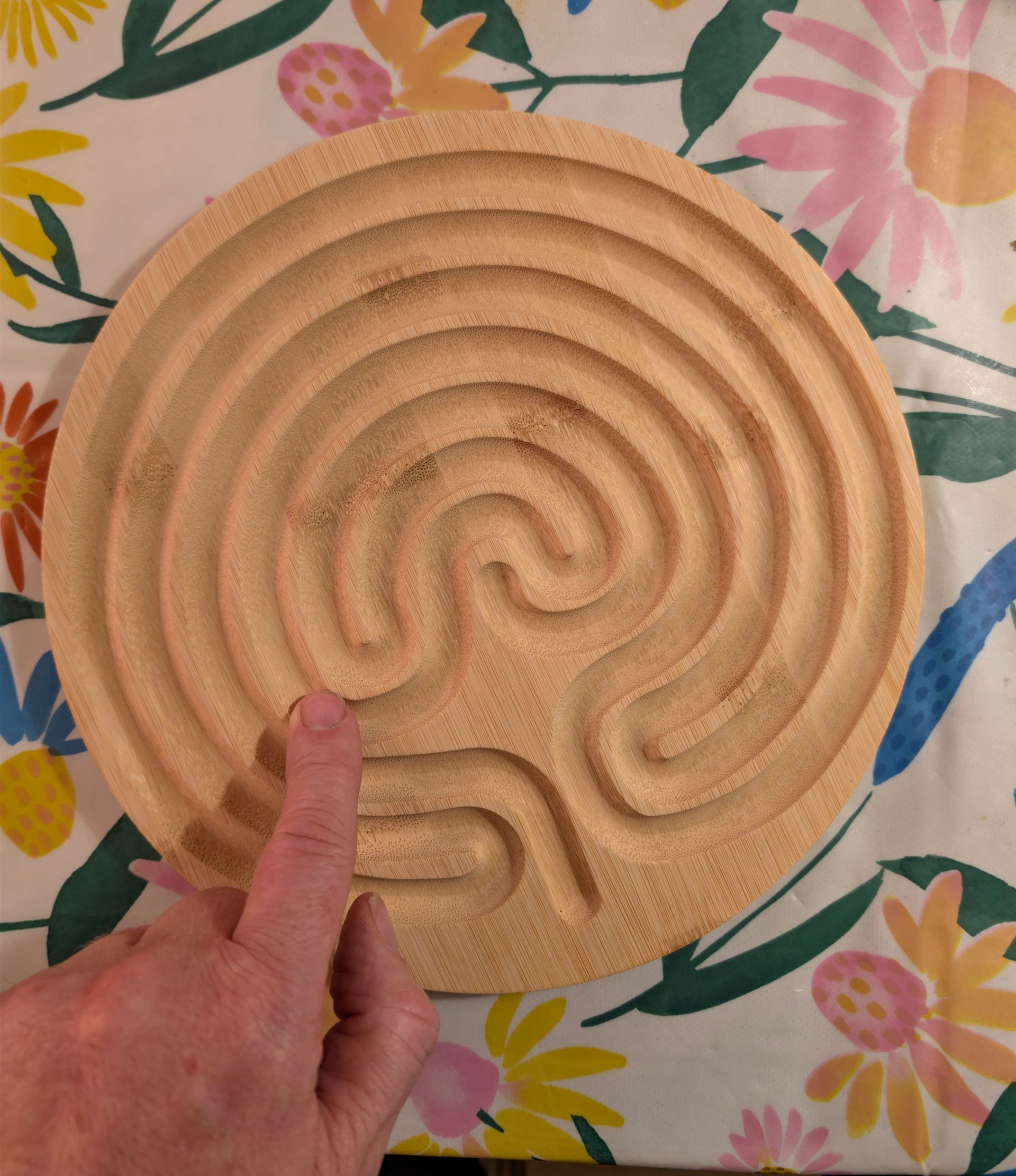 Houten labyrint, vinger wijst naar het begin. Lichtgekleurd hout, cirkelvormig patroon op een bloemenkleed.