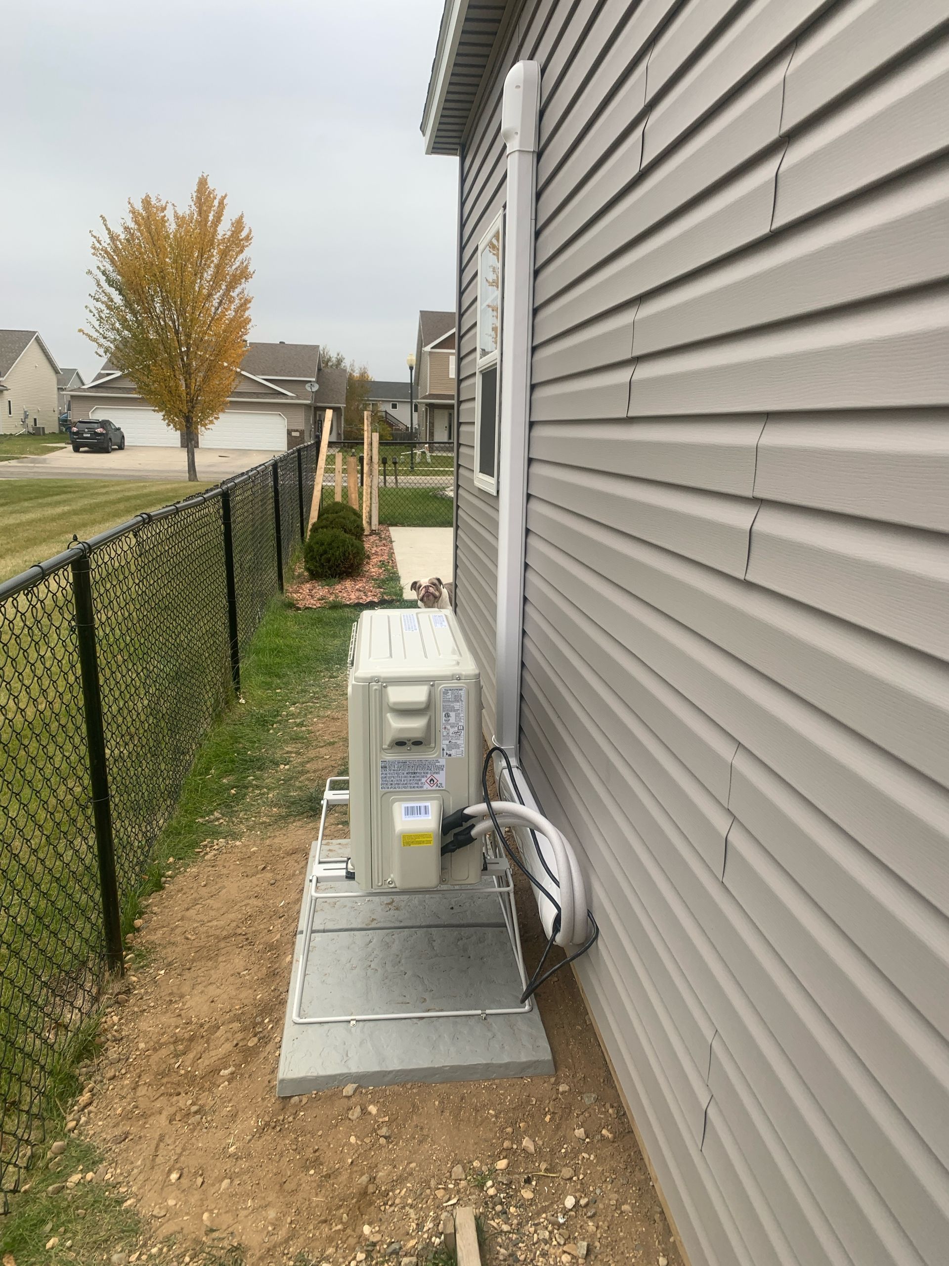 An air conditioning unit sits on a metal base next to a house with gray siding and a black fence.