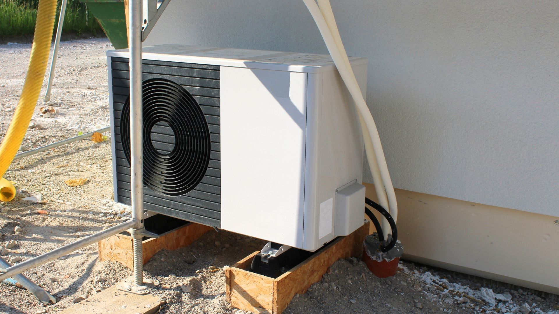 An outdoor air conditioning unit, white, mounted on wood supports next to a building.