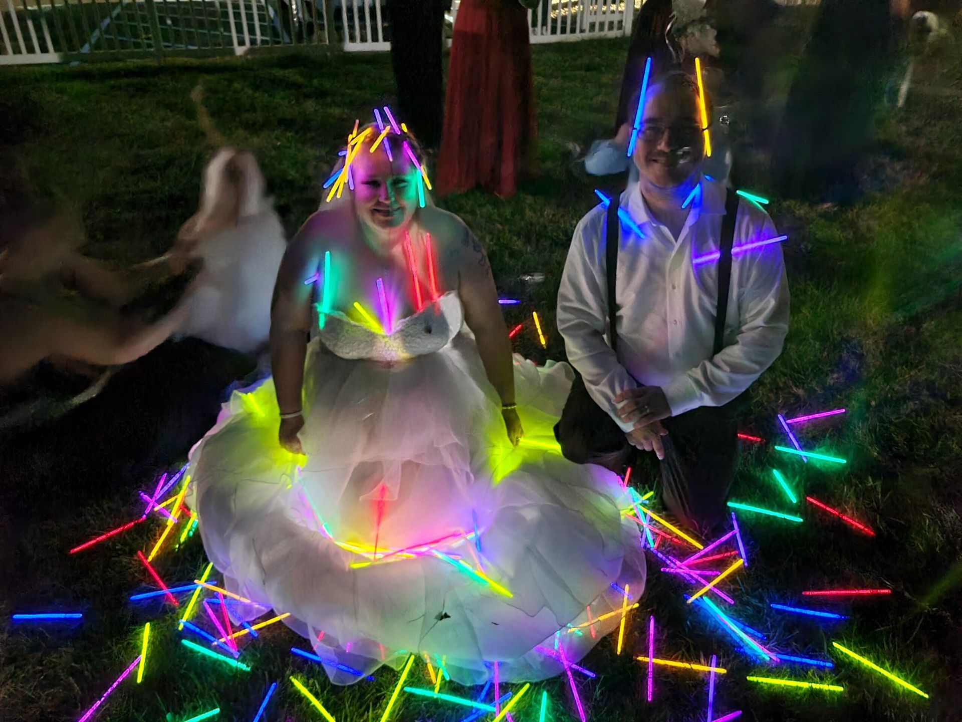 A bride and groom are sitting on the grass surrounded by glow in the dark sticks.