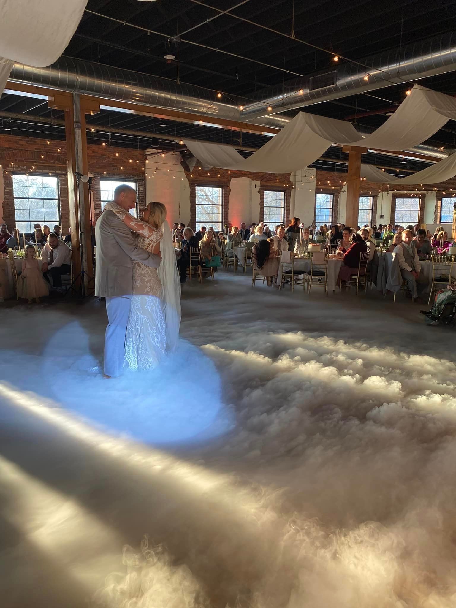 A bride and groom are dancing in a large room with smoke coming out of the ceiling.