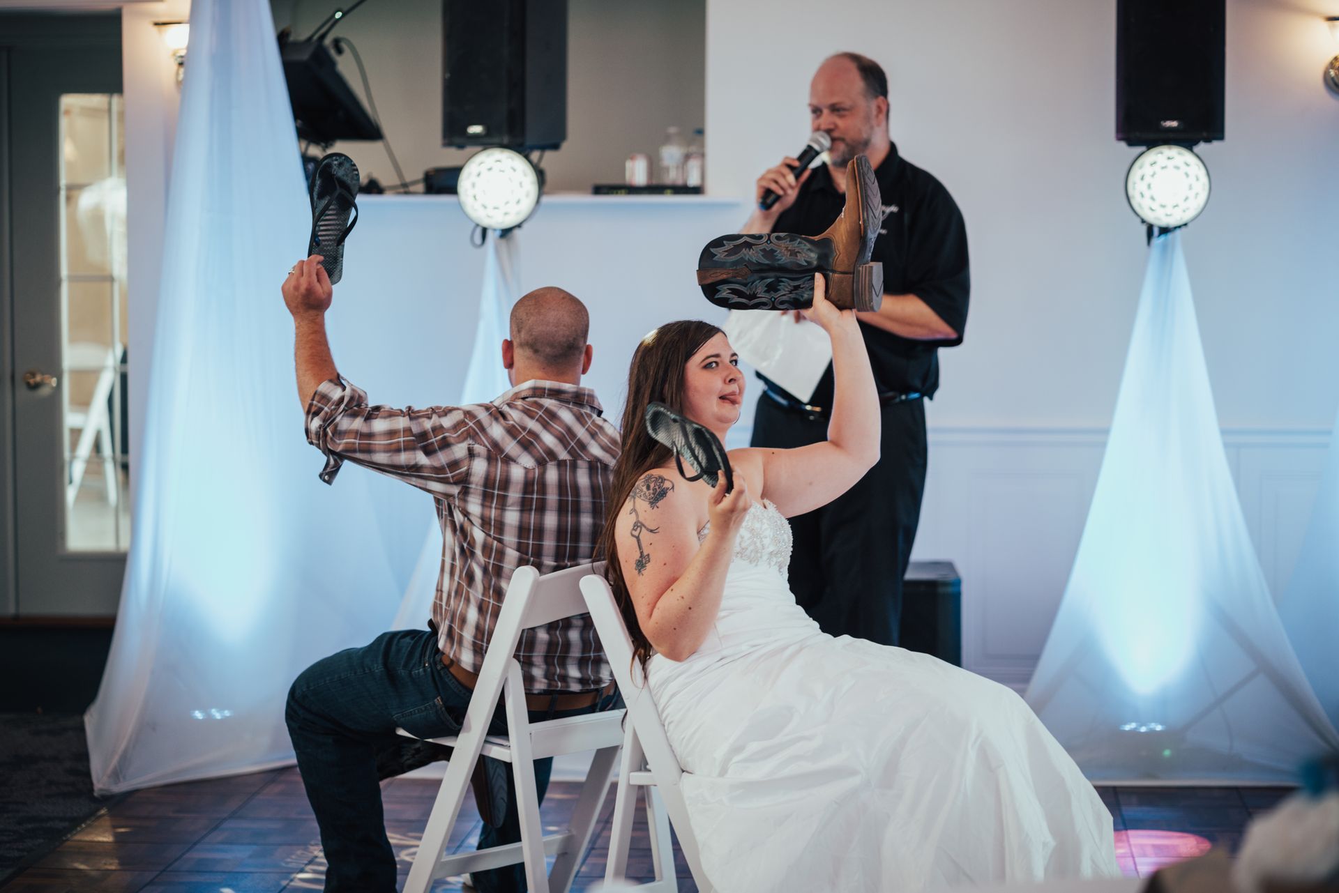 A bride and groom are sitting in chairs at a wedding reception while a man sings into a microphone.