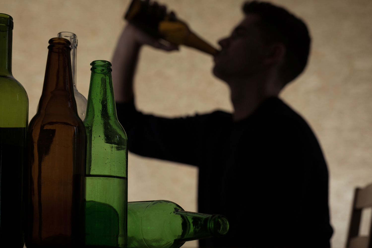 Silhouette of person drinking from a bottle, with several empty bottles on a table in the foreground.