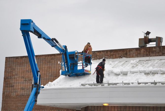 Workers in a blue lift clearing snow from a rooftop beside a brick building