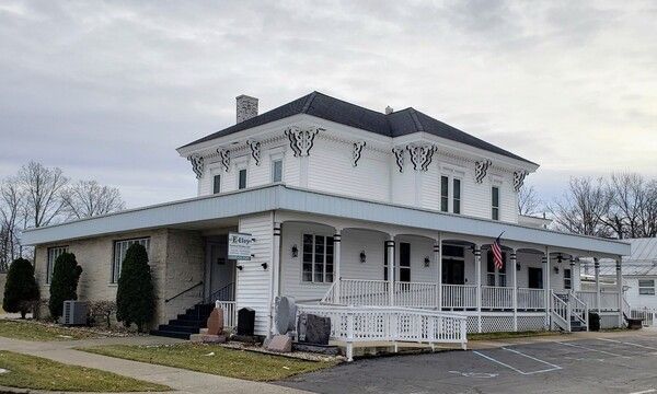 White, two-story building with a porch and flat roof. An American flag hangs. Cloudy sky background.