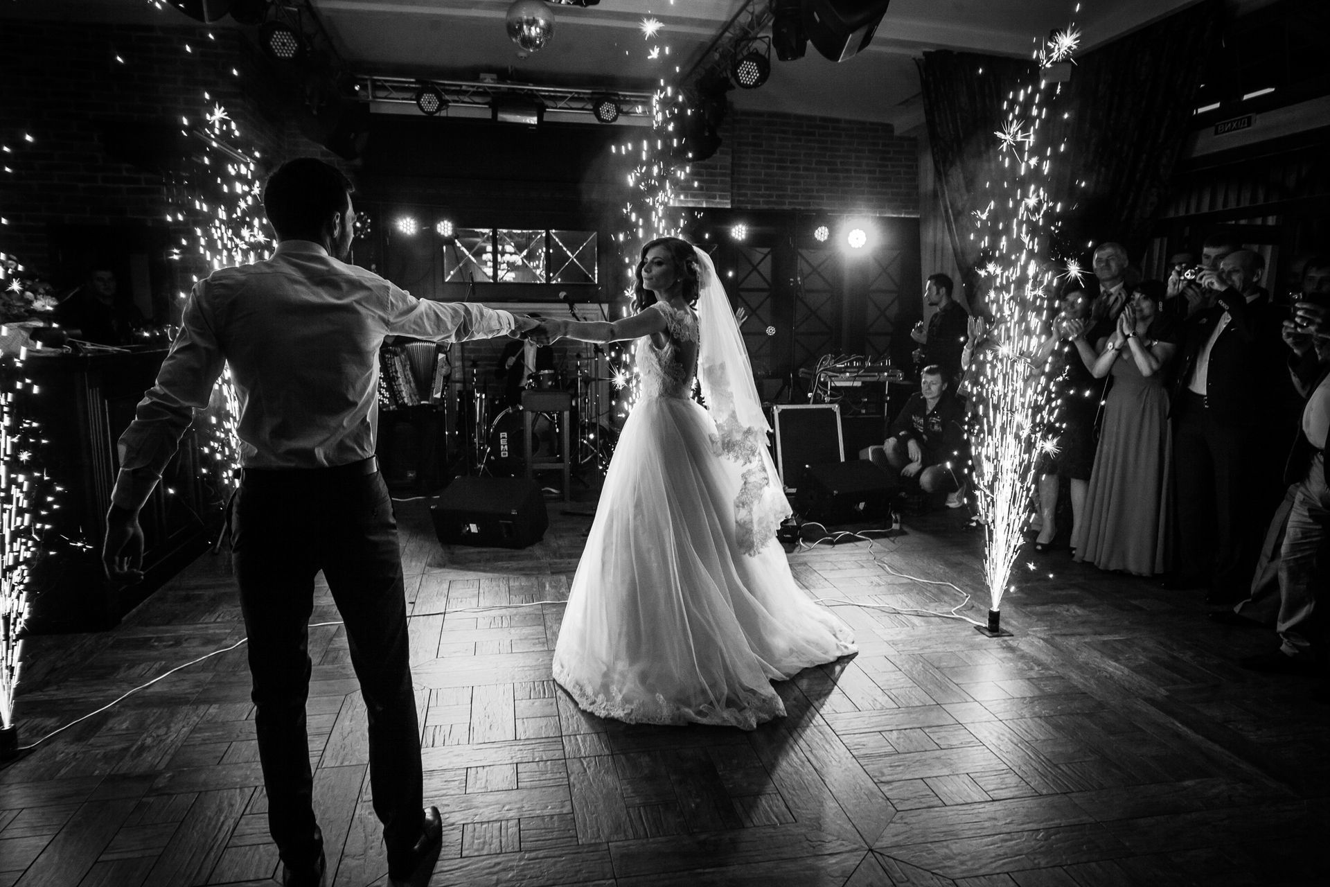 A bride and groom share their first dance on a wooden dance floor surrounded by pyrotechnic sparks in a dimly lit venue.