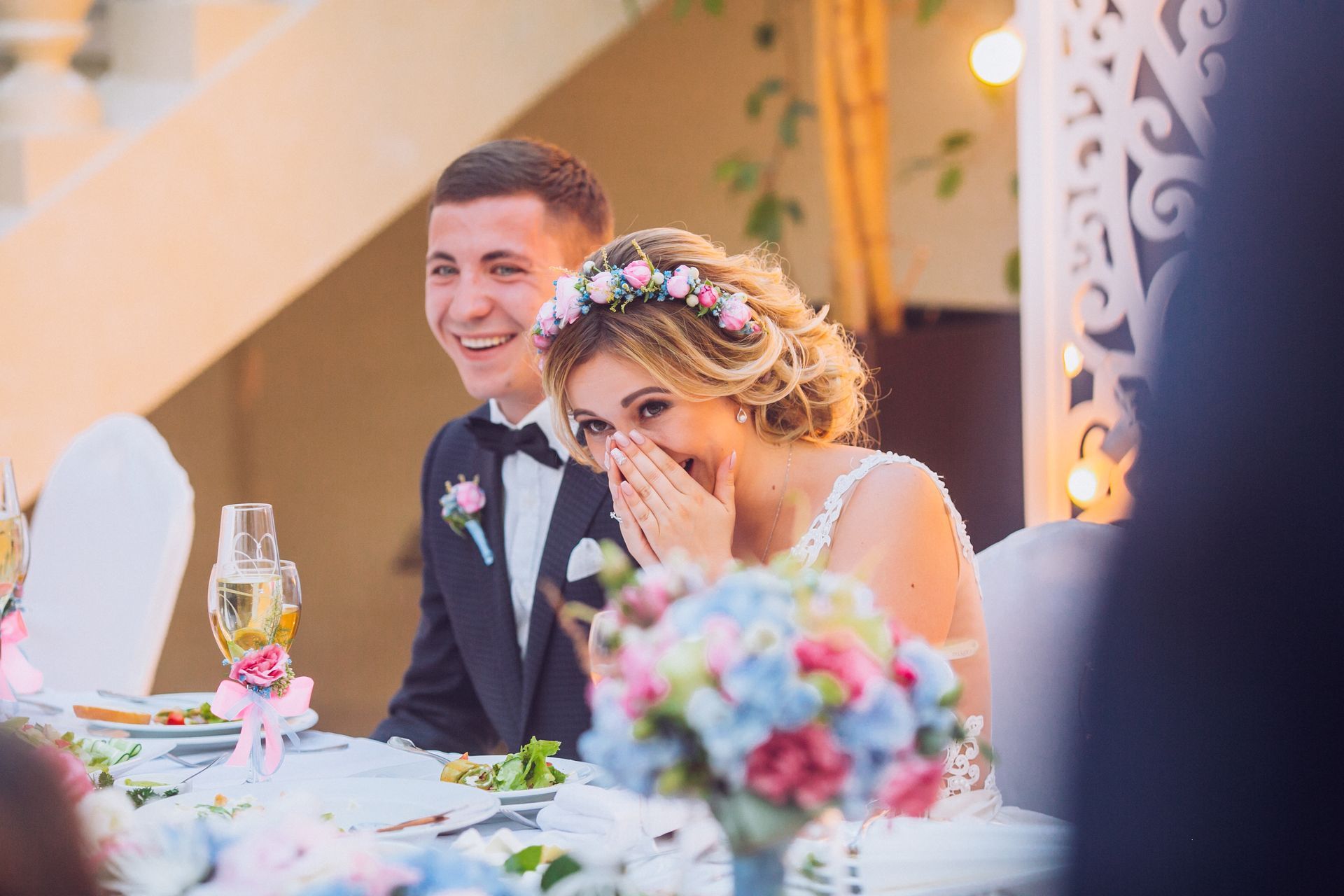 A bride in a floral crown laughs while covering her mouth, sitting next to a smiling groom at a wedding reception table.