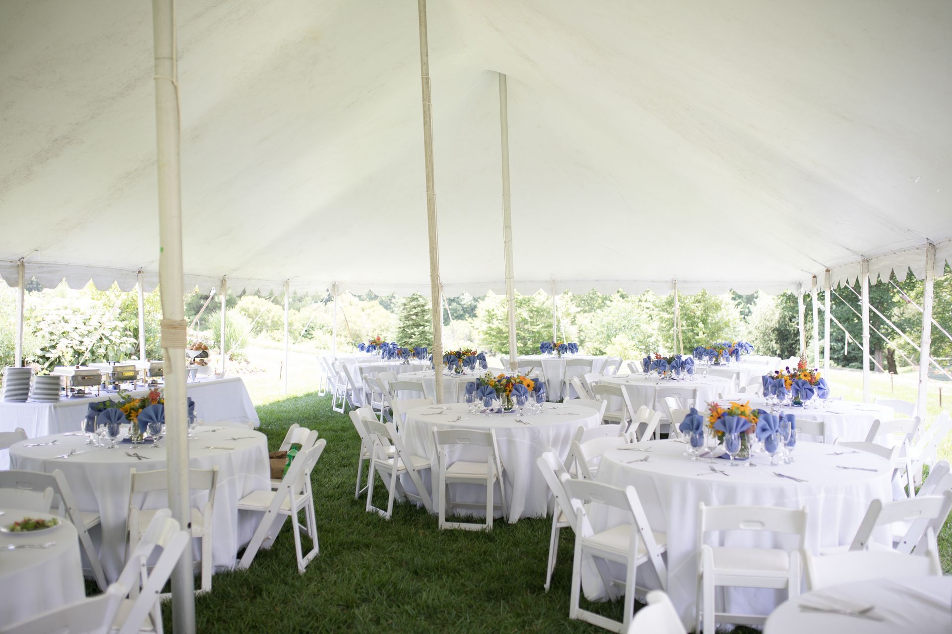 A white tent set up for an outdoor event with round tables covered in white linens and white chairs on a grassy lawn.