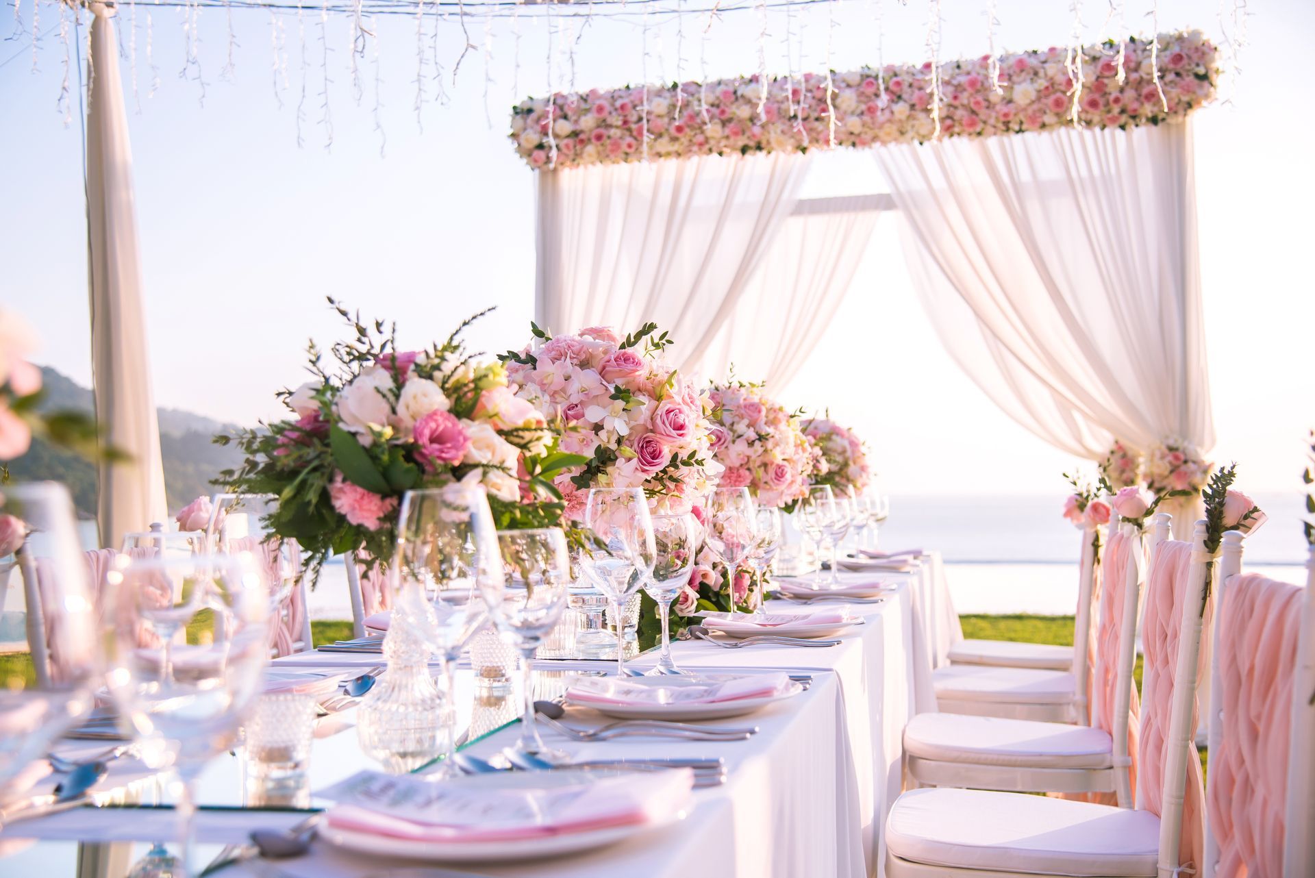 A beach wedding reception setup with floral centerpieces, white table linens, and a decorated arch overlooking the ocean.