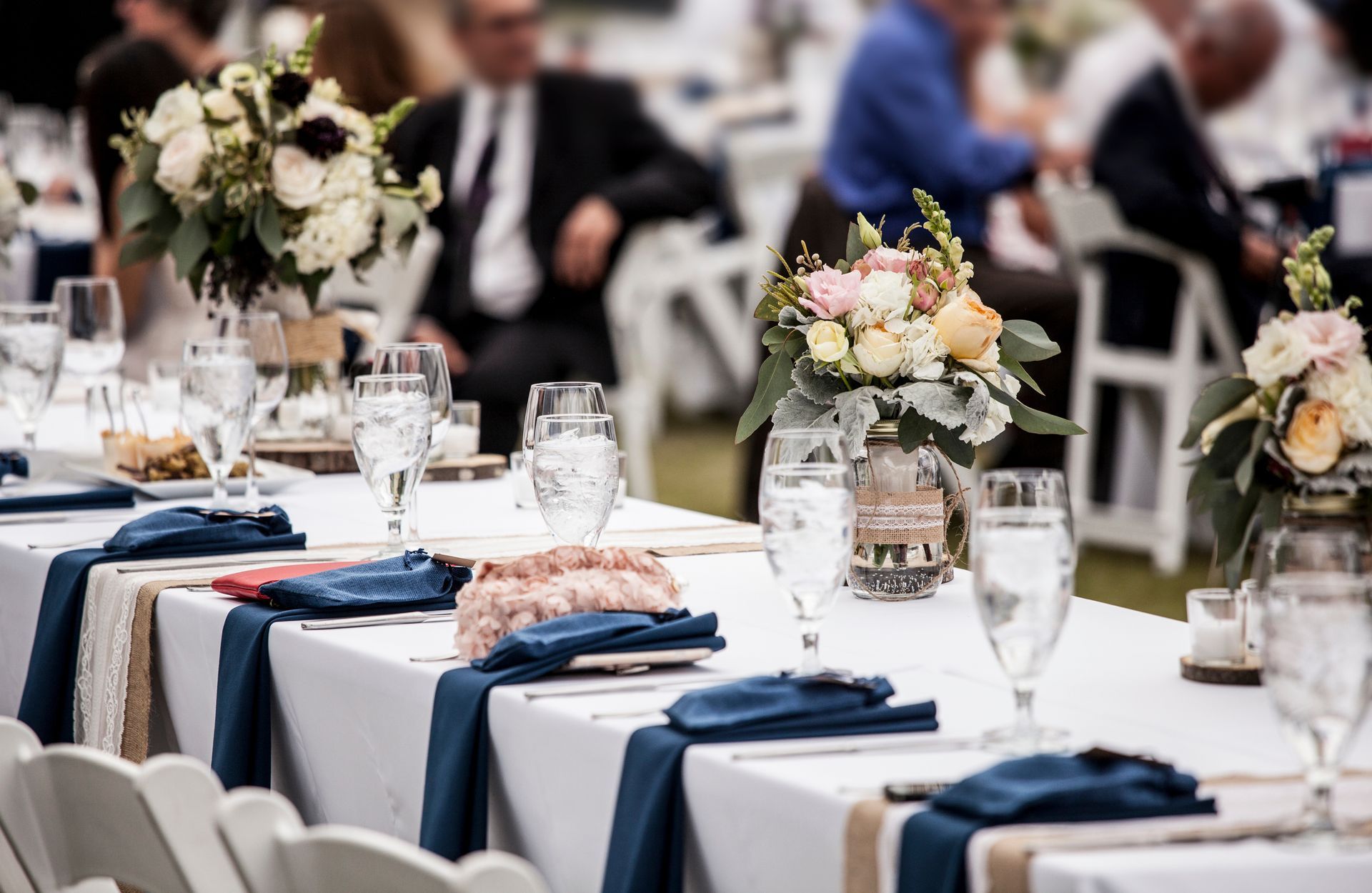 A long banquet table set with white tablecloths, navy napkins, glassware, and floral centerpieces at an event.