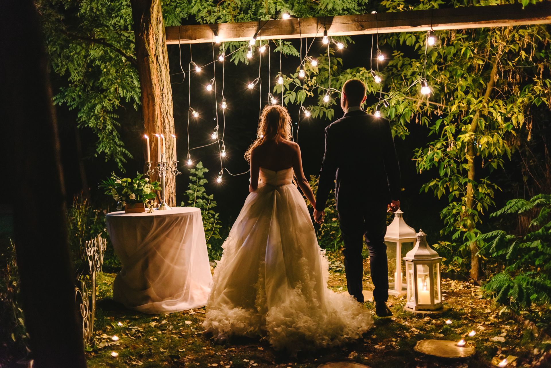 A couple standing hand-in-hand in a garden at night, illuminated by string lights and lanterns.