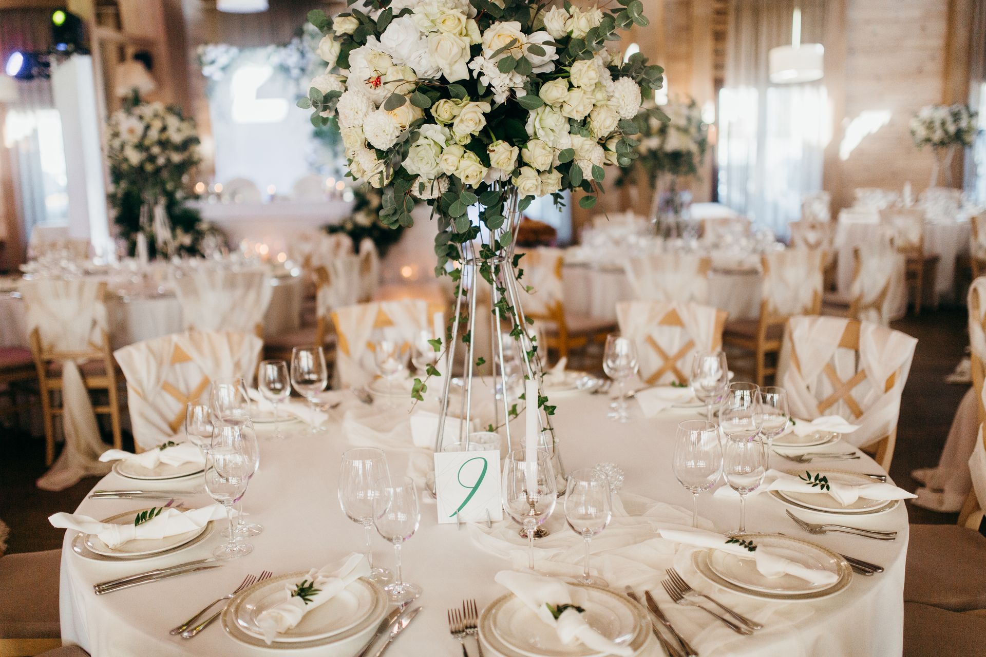 Elegant wedding reception table with a white tablecloth, gold-accented chairs, and a large white floral centerpiece.