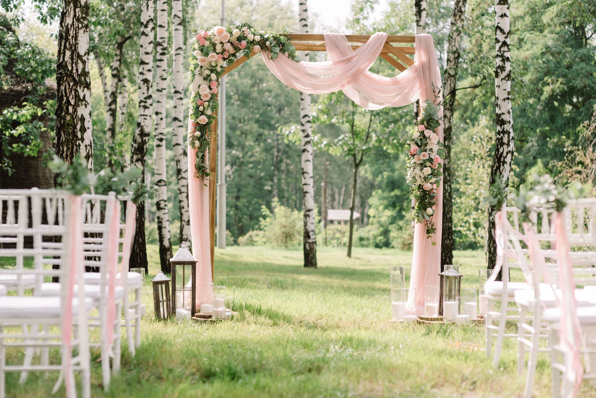 Outdoor wedding aisle with white chairs, pink drapes, and floral garlands under trees