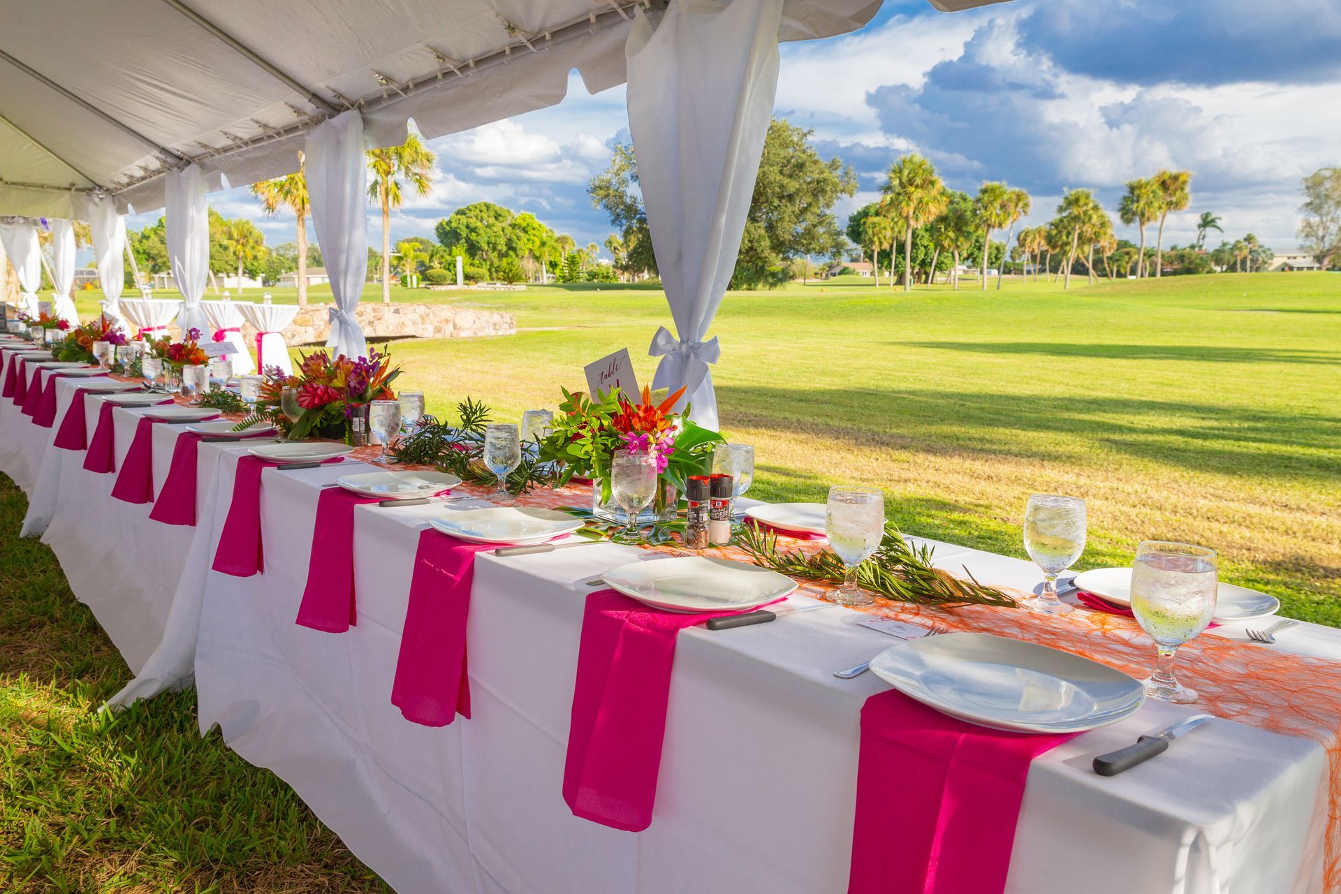 Long banquet table with white tablecloths and hot pink runners under a tent overlooking a sunny green golf course.