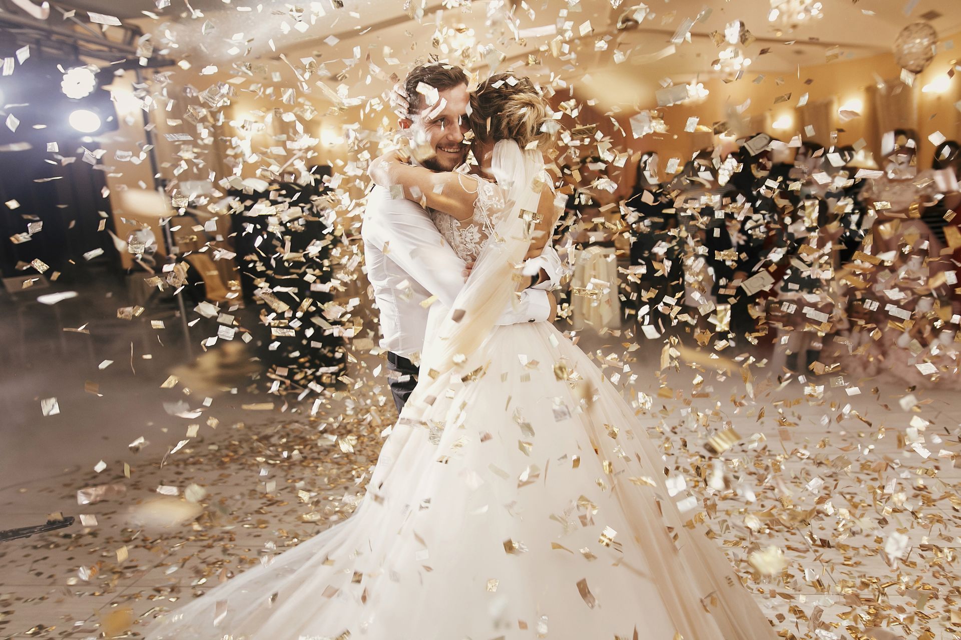 A bride and groom embrace on a dance floor surrounded by falling gold confetti.