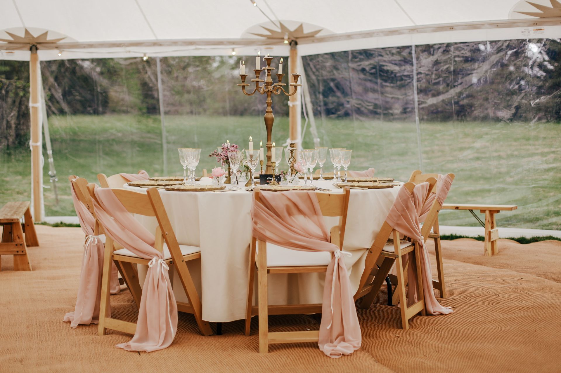 A circular wedding table with white linens, gold candelabra, and wooden chairs draped in soft pink fabric under a tent.