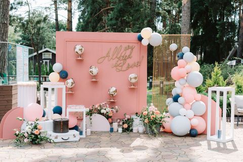 A pink wedding backdrop with globes, floral arrangements, and a balloon arch, set outdoors in a wooded area. A pink wedding backdrop with globes, floral arrangements, and a balloon arch, set outdoors in a wooded area.
